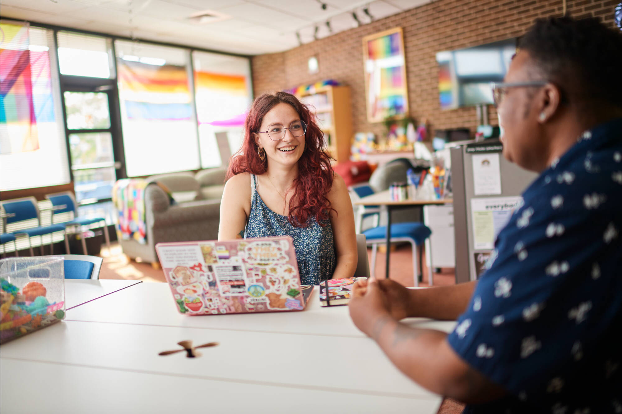 Two individuals engage in conversation at a table in a brightly lit, colorful room with rainbow flags and creative decor.